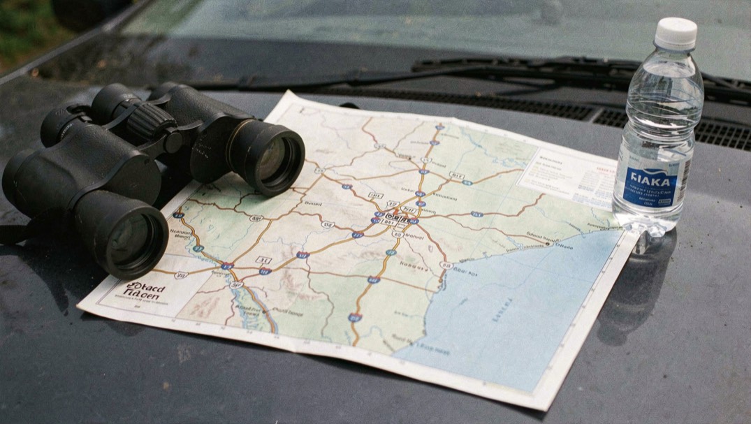 Map spread on a car hood beside binoculars and a water bottle showing southwest highways between tiny numbered site markers