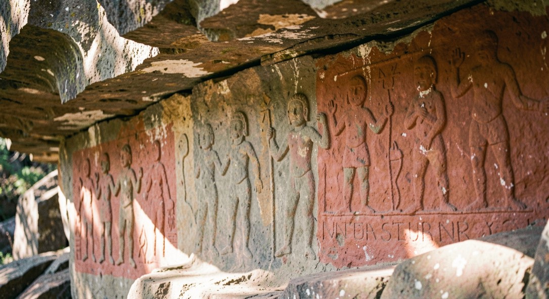Deeply patinated petroglyph figures beside a painted red ochre panel under a rock overhang lit by reflected morning light